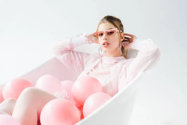 Mujer joven con estilo en gafas de sol tumbado en la bañera con globos de aire de color rosa en blanco — Stock Photo