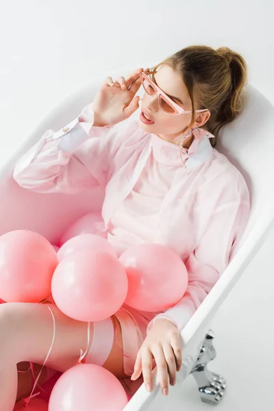 Overhead view of attractive girl touching sunglasses while lying in bathtub with pink air balloons on white — Stock Photo