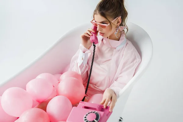 Приваблива дівчина в сонцезахисних окулярах говорить на ретро телефоні, лежачи у ванній з рожевими повітряними кулями на білому — стокове фото