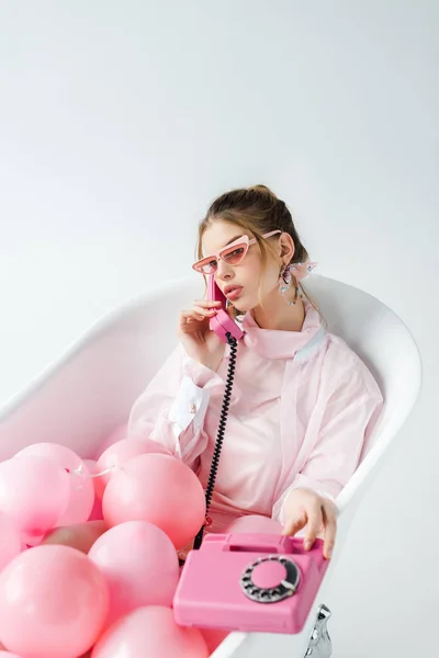 Girl in sunglasses talking on retro phone while lying in bathtub with pink air balloons on white — Stock Photo