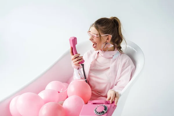 Girl in sunglasses screaming at pink retro phone while lying in bathtub with air balloons on white — Stock Photo