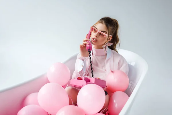 Mujer joven en gafas de sol hablando en el teléfono retro mientras se encuentra en la bañera con globos de aire de color rosa en blanco - foto de stock