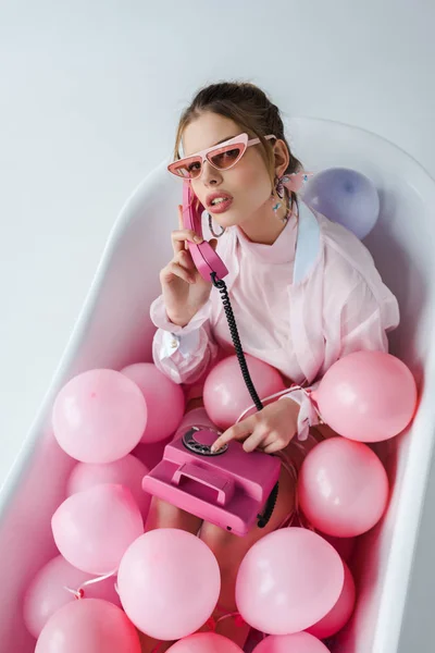 Vista aérea de mujer joven en gafas de sol hablando en el teléfono retro mientras que miente en la bañera con globos de aire rosados en blanco — Stock Photo