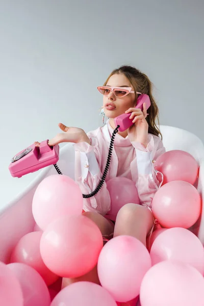 Woman in sunglasses talking on retro phone while lying in bathtub with pink air balloons on white — Stock Photo