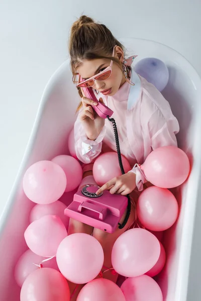 Вид зверху дівчини в сонцезахисних окулярах розмовляє на рожевому ретро телефоні, лежачи у ванній з повітряними кулями на білому — стокове фото