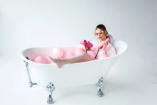 Дівчина в сонцезахисних окулярах дивиться на рожевий ретро телефон, лежачи у ванній з повітряними кулями на білому — стокове фото