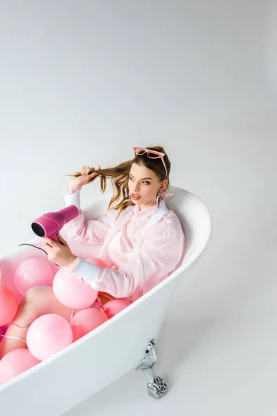 Привлекательная девушка с феном во время лежания в ванне с розовыми воздушными шарами на сером — стоковое фото