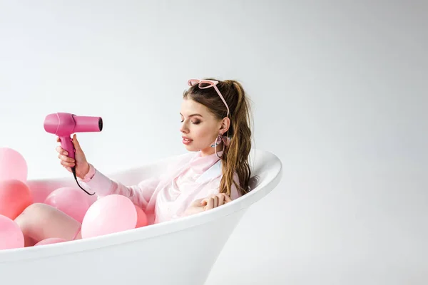 Hübsches Mädchen mit Haartrockner in der Badewanne liegend mit rosa Luftballons auf weiß — Stockfoto