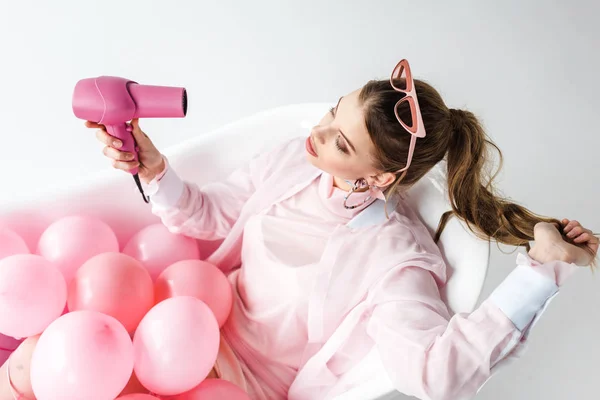 Overhead view of girl using hair dryer while lying in bathtub with pink air balloons on white — Stock Photo