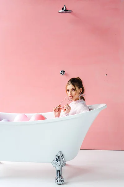 Bonita chica sosteniendo gafas de sol mientras está acostado en la bañera en rosa - foto de stock