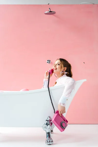 Happy girl talking on retro phone while lying in bathtub on pink — Stock Photo