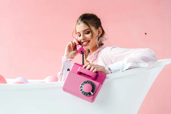 Cheerful girl talking on retro phone while lying in bathtub on pink — Stock Photo