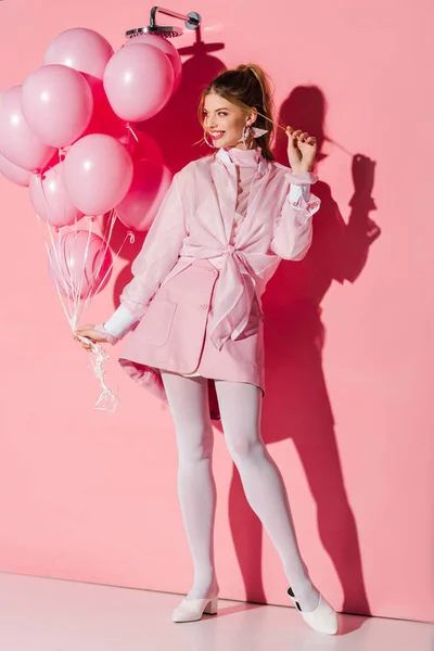 Fröhliche junge Frau hält Luftballons in der Hand, während sie die Haare auf rosa berührt — Stockfoto