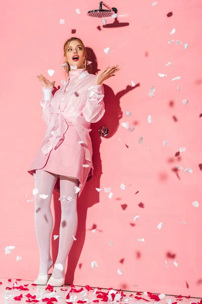 Attractive surprised young woman standing near rose petals on pink — Stock Photo