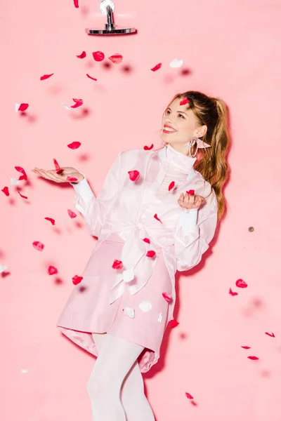 Attractive cheerful young woman standing near rose petals on pink — Stock Photo