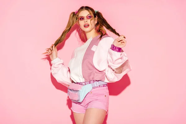 Удивлена стильная девушка в солнцезащитных очках и талии мешок касаясь волос на розовый — стоковое фото