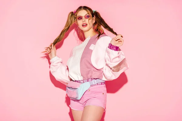 Surprised stylish girl in sunglasses and waist bag touching hair on pink — Stock Photo