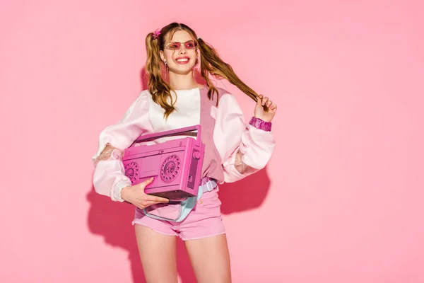 Happy stylish girl in sunglasses holding boombox and touching hair on pink — Stock Photo