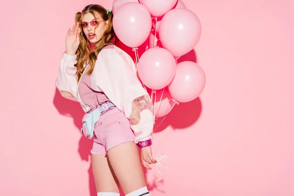 Überraschte junge Frau mit Sonnenbrille und Luftballons auf rosa — Stockfoto