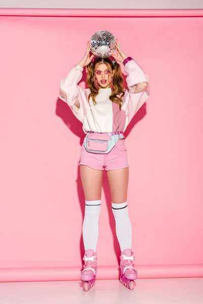 Attractive girl in sunglasses holding shiny disco ball above head while standing in roller-skates on pink — Stock Photo