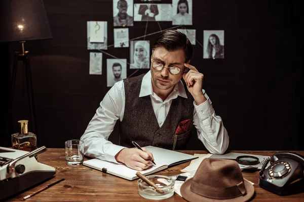 Detective pensativo en vasos sentados a la mesa y escribiendo en cuaderno - foto de stock