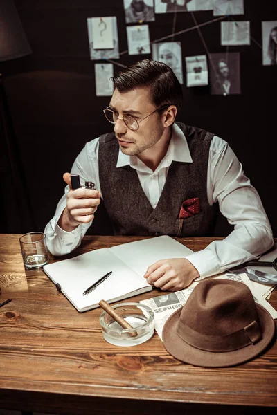 Pensive detective in glasses sitting at table and holding lighter in office — Stock Photo