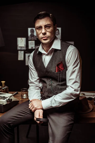 Detective in glasses sitting on table and holding magnifier in dark office — Stock Photo