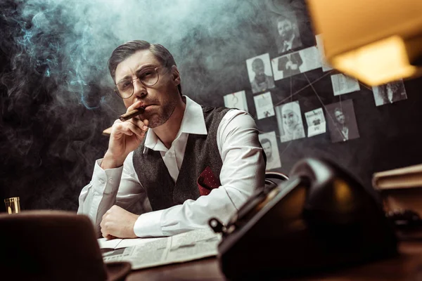 Vista a basso angolo di detective pensoso in occhiali fumare sigaro in ufficio — Foto stock