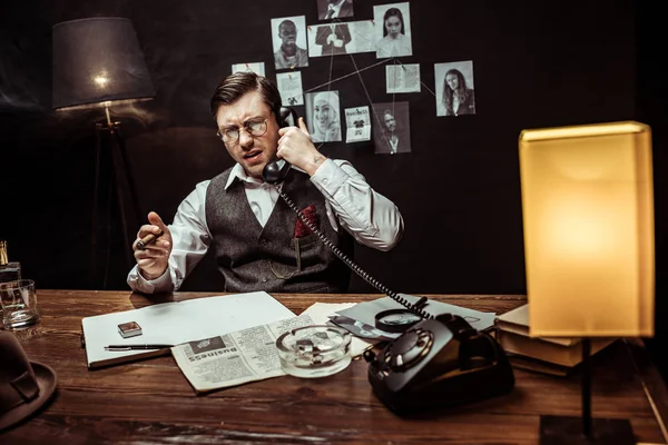 Irritierter Detektiv mit Brille, Zigarre in der Hand und Telefongespräch im dunklen Büro — Stockfoto