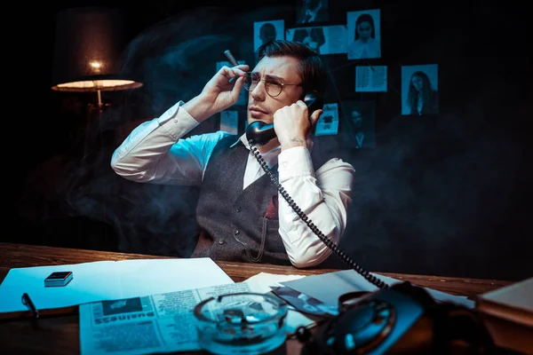 Детектив в очках, держит сигару и разговаривает по телефону в темном кабинете — стоковое фото