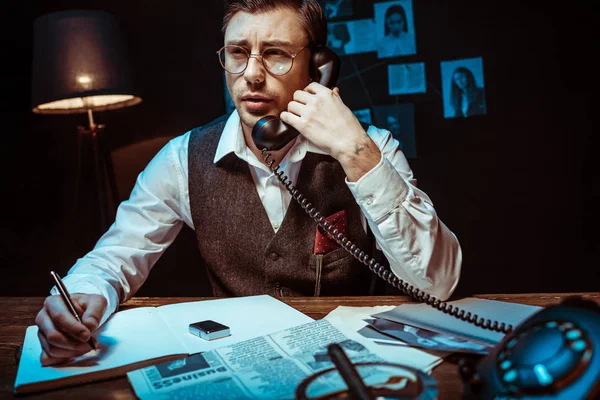 Концентрований детектив в окулярах розмовляє по телефону і пише в блокноті в офісі — стокове фото