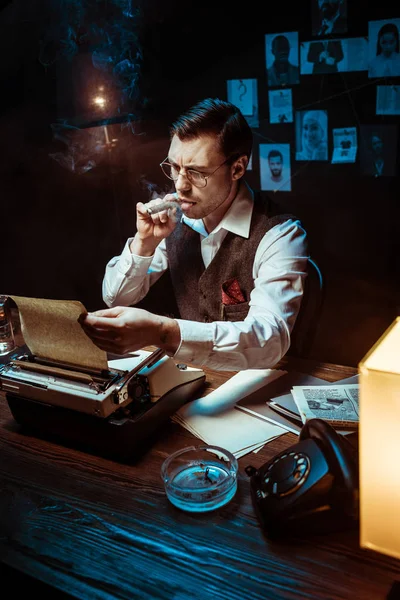 Détective dans des lunettes fumant cigare tout en utilisant une machine à écrire dans le bureau sombre — Photo de stock