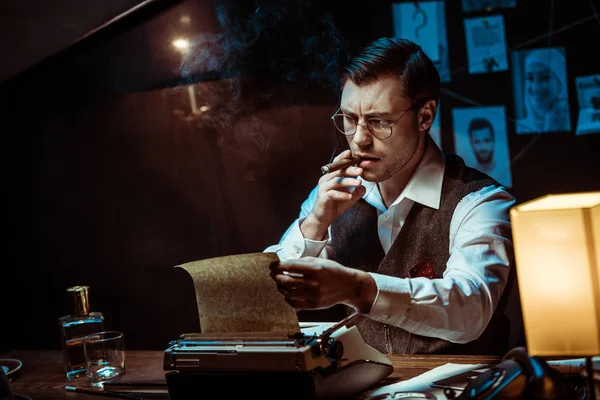 Detective in glasses smoking cigar while using typewriter in dark office — Stock Photo