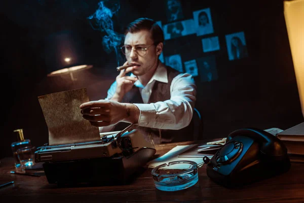 Detective en gafas fumar cigarro mientras usa la máquina de escribir en la oficina oscura - foto de stock