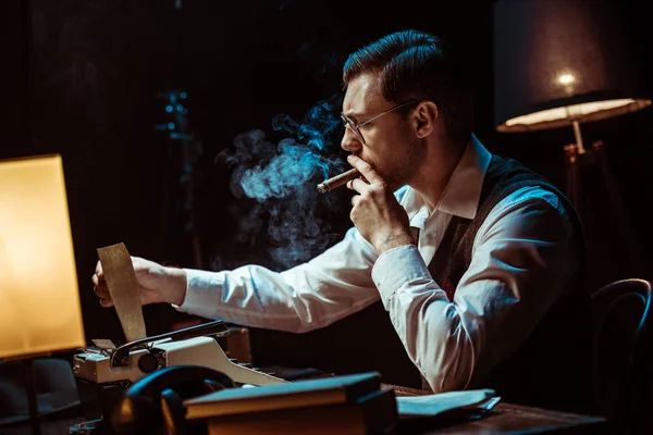 Détective dans des lunettes fumant cigare tout en utilisant une machine à écrire dans le bureau sombre — Photo de stock