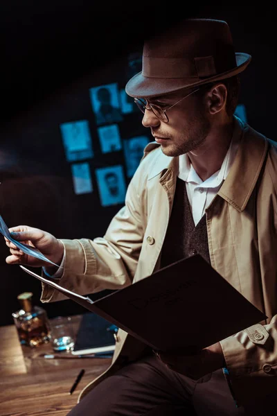 Detective in trench coat and hat holding dossier and looking at photo — Stock Photo