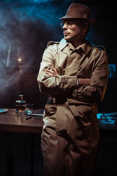 Detective in trench coat and hat standing with crossed arms in office — Stock Photo