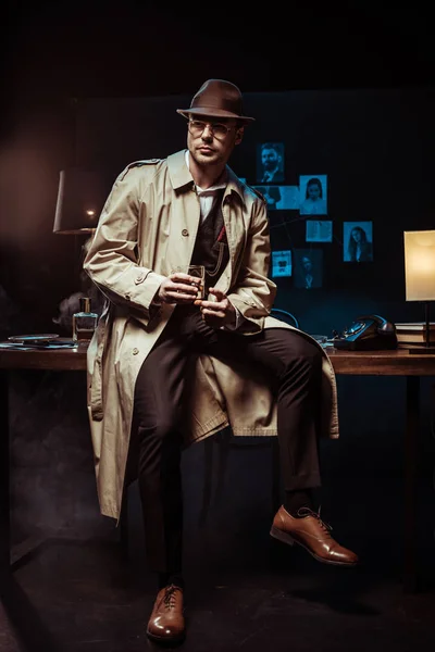 Détective en chapeau et trench coat assis sur la table avec verre de cognac — Photo de stock