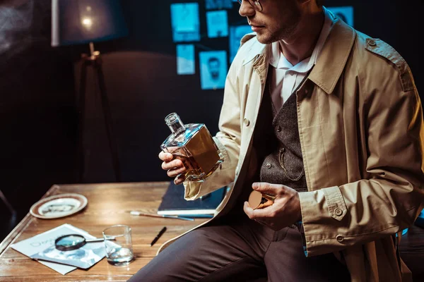 Vista recortada del detective en gabardina sentado en la mesa y sosteniendo la botella de coñac - foto de stock