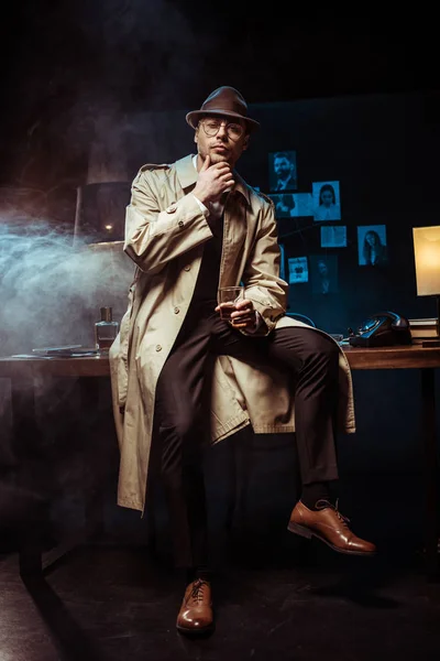 Detective in trench coat and hat sitting on table and holding glass of cognac — Stock Photo