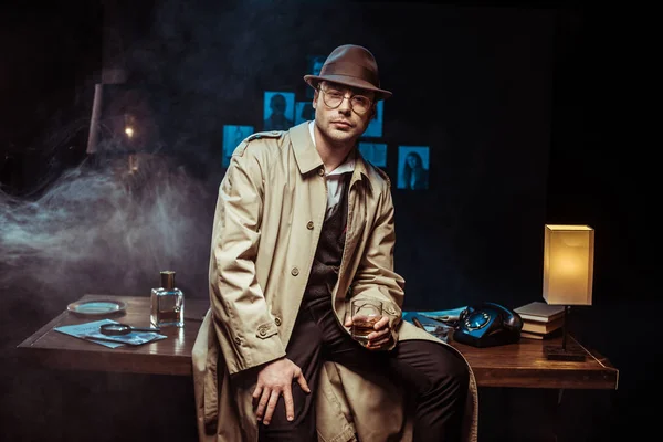 Detektiv in Trenchcoat und Hut sitzt auf dem Tisch und hält ein Glas Cognac — Stockfoto
