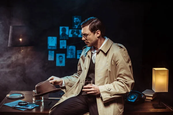 Detektiv im Trenchcoat sitzt auf Tisch mit Hut und Glas Cognac — Stockfoto