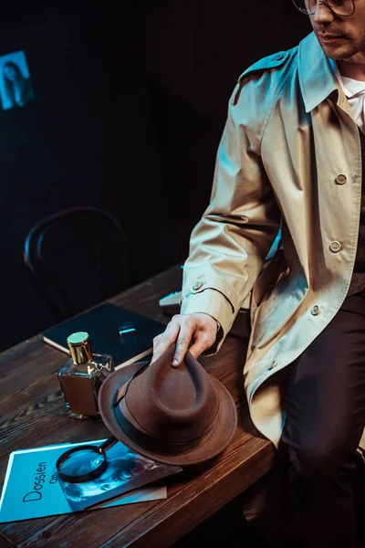 Cropped view of detective in trench coat sitting on table and holding hat — Stock Photo