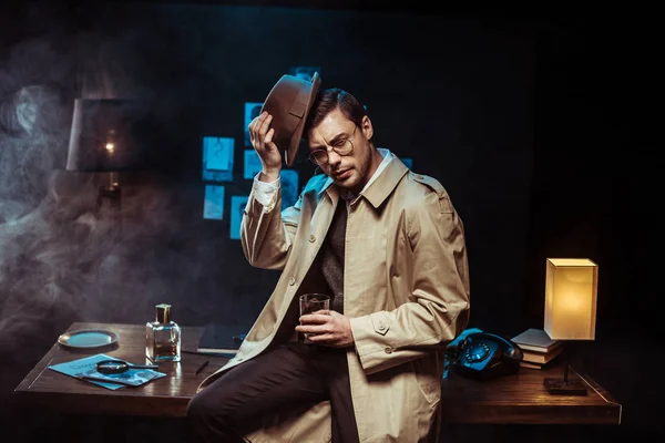Nachdenklicher Detektiv setzt Hut auf und hält Glas Cognac in der Hand — Stockfoto