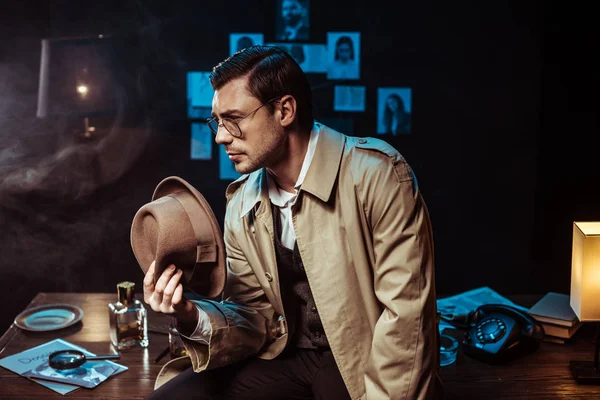 Pensive detective in glasses and trench coat sitting on table and holding hat — Stock Photo