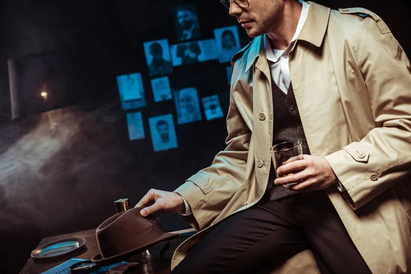 Partial view of detective in trench coat holding hat and glass of cognac — Stock Photo