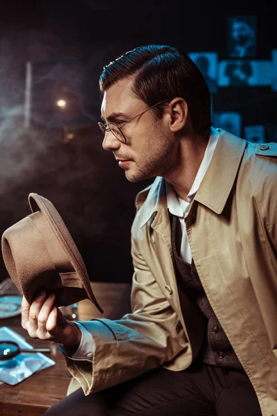Nachdenklicher Detektiv in Brille und Trenchcoat sitzt am Tisch und hält Hut — Stockfoto