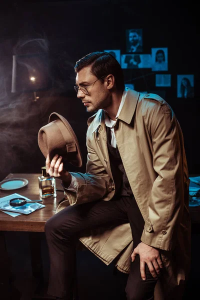 Pensive detective in glasses and trench coat sitting on table and holding hat — Stock Photo