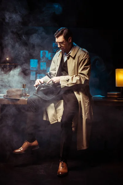 Детектив в траншеї пальто сидить на столі і використовує телефон в темному офісі — стокове фото
