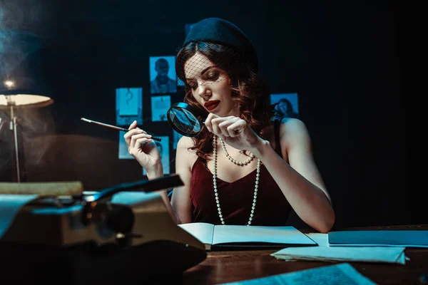 Attrayant femme dans le chapeau fascinateur tenant embout buccal et loupe dans le bureau sombre — Photo de stock